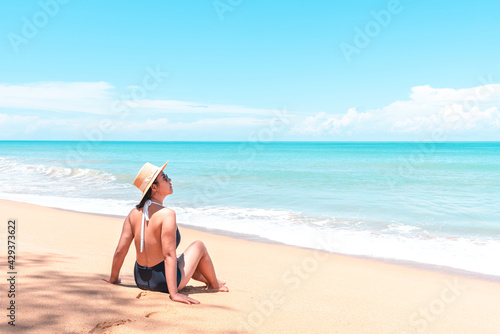 Back view of asian traveler woman in swimsuit and straw hat to sitting on the beach to looking view the blue horizon
