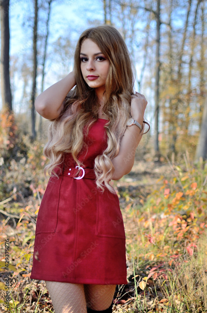 Warm autumn in Poland. Young woman with red dress surrounded by trees. Woman with blonde hair extension.