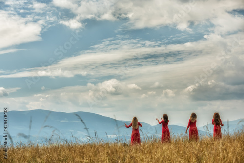 Girls standing with their backs with outstretched arms in red dresses on the edges of a meadow overlooking the mountains.