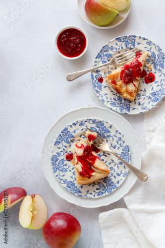 Two pieces of apple tart with raspberries jam on blue plates and some apples, shot from above