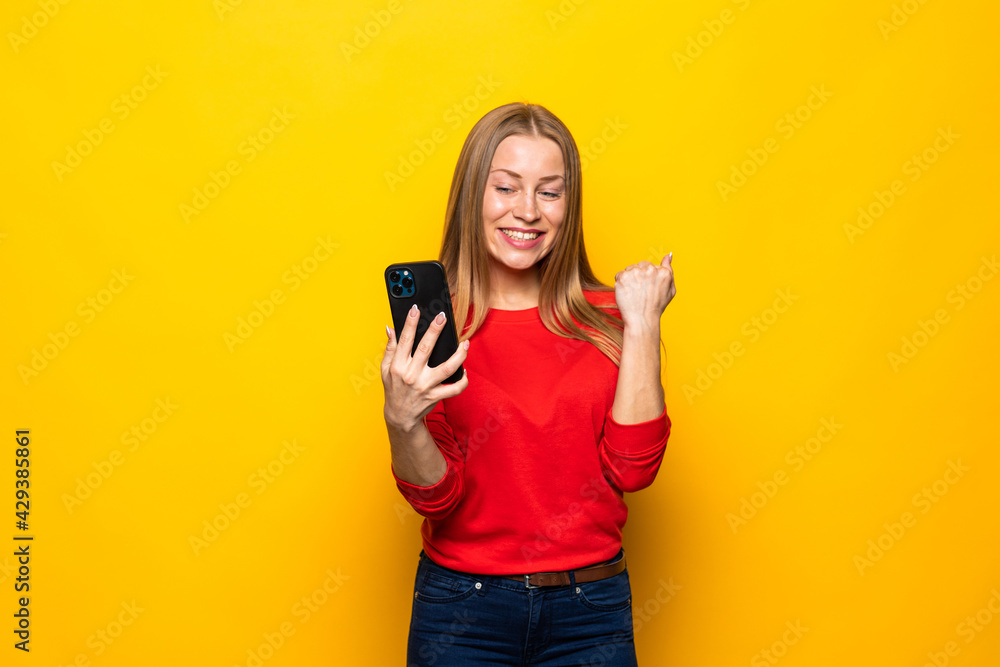 Young woman use smart phone celebrate social network lottery victory raise fists scream isolated on yellow background Young woman use smart phone celebrate social network lottery victory raise fists scream isolated on yellow background