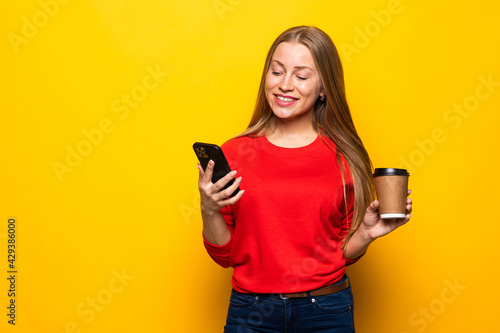 Beautiful young woman drinking coffee using mobile phone posing isolated over yellow wall background