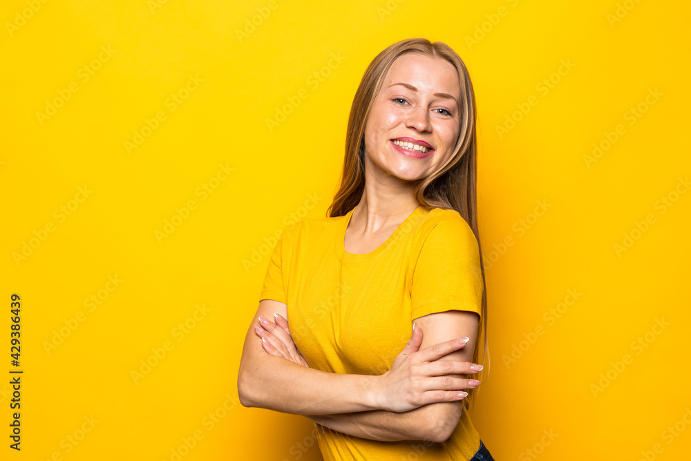 Obraz premium Portrait of a smiling blonde young woman standing with arms folded isolated over yellow background