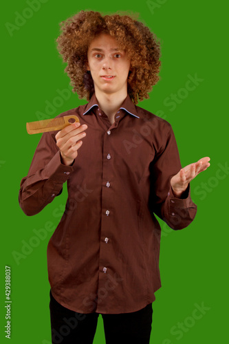 A long-haired curly-haired guy in a brown shirt on a green background uses a wooden comb. Emotions before a haircut in a hairdresser