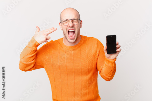 bald man with a smartphone looking unhappy and stressed, suicide gesture making gun sign with hand, pointing to head photo