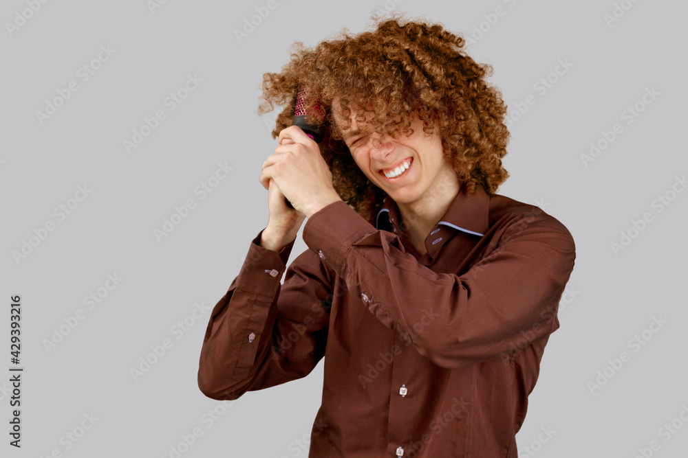A long-haired curly-haired guy in a brown shirt on a gray background uses a metal round comb. Emotions before a haircut in a hairdresser. Pain from combing A long-haired curly-haired guy in a brown shirt on a gray background uses a metal round comb. Emotions before a haircut in a hairdresser. Pain from combing