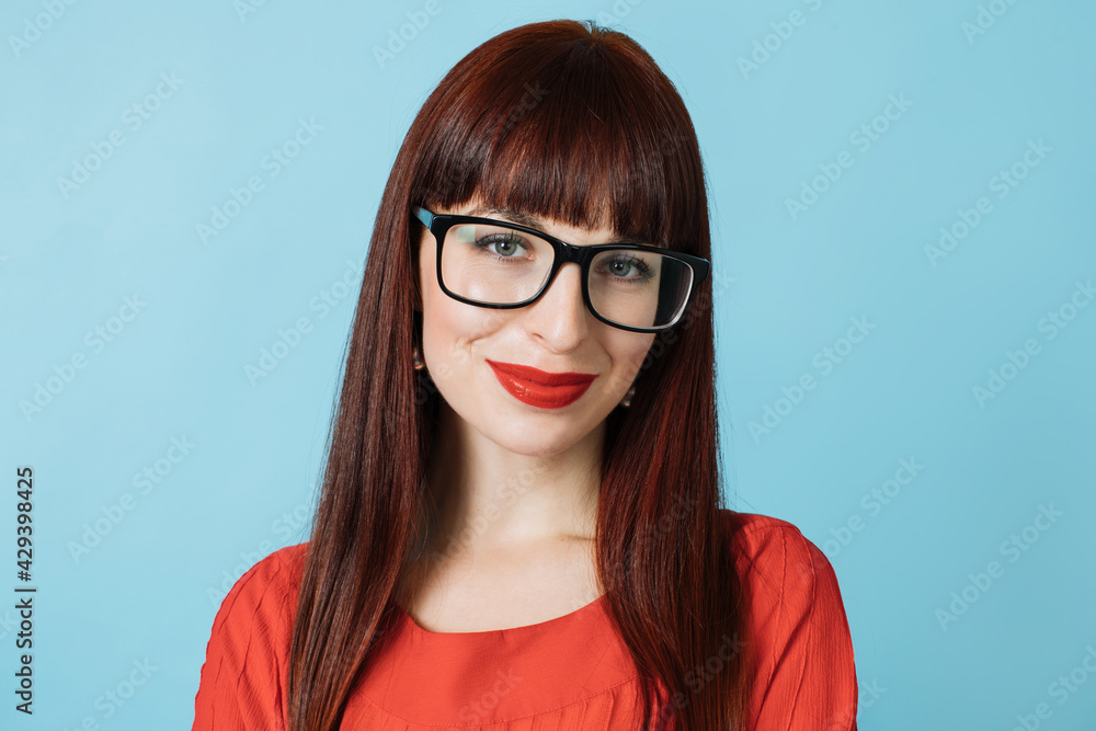 Pleasant beautiful red haired woman in eyeglasses, wearing red shirt, red lipstick, smiling at camera on blue background. Close up portrait with copy space