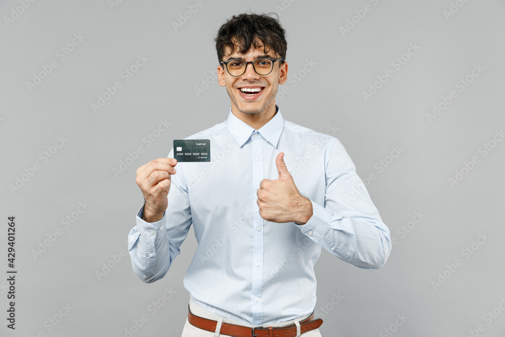 Young rich smiling employee business latin man corporate lawyer 20s wearing classic white shirt glasses hold in hand credit bank card show thumb up gesture isolated on grey background studio portrait Young rich smiling employee business latin man corporate lawyer 20s wearing classic white shirt glasses hold in hand credit bank card show thumb up gesture isolated on grey background studio portrait