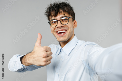 Close up young smiling happy employee business latin man 20s corporate lawyer in classic white shirt glasses doing selfie shot on mobile phone show thumb up gesture isolated on grey background studio