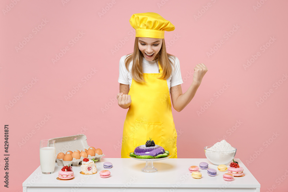Teen fun girl chef cook confectioner baker in yellow apron cap at table do winner gesture clench fist celebrate isolated on pastel pink background Mousse cake cooking food process workshop concept. Teen fun girl chef cook confectioner baker in yellow apron cap at table do winner gesture clench fist celebrate isolated on pastel pink background Mousse cake cooking food process workshop concept.