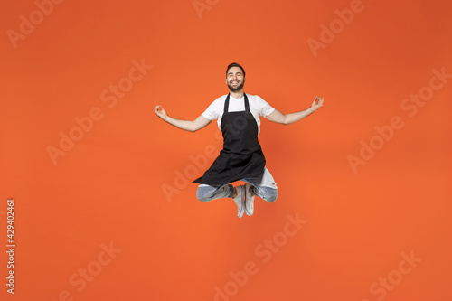 Full length young man 20s barista bartender barman employee in black apron white t-shirt work in coffee shop jump high do yoga om gesture isolated on orange background. Small business startup concept. © ViDi Studio