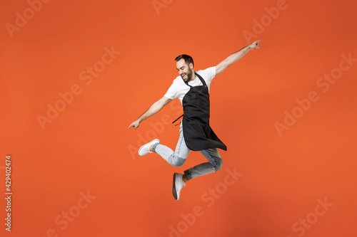 Full length young fun man barista bartender barman employee in black apron white t-shirt work in coffee shop jump high outstretched hands isolated on orange background. Small business startup concept.