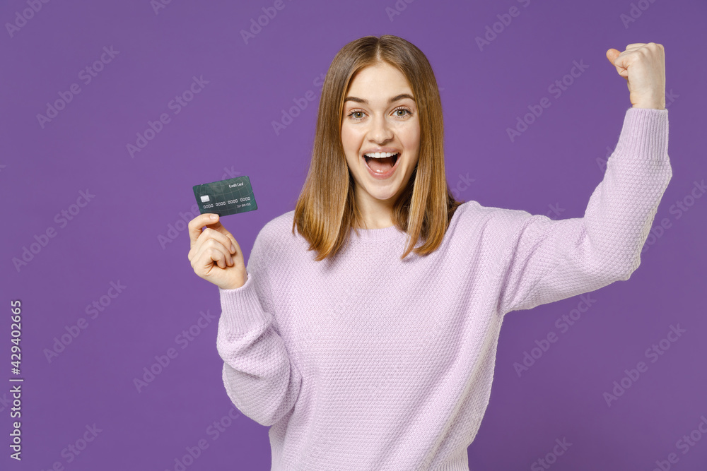 Young overjoyed rich happy student woman 20s wearing purple knitted sweater hold credit bank card do winner gesture clench fist isolated on violet background studio portrait. People lifestyle concept. Young overjoyed rich happy student woman 20s wearing purple knitted sweater hold credit bank card do winner gesture clench fist isolated on violet background studio portrait. People lifestyle concept.