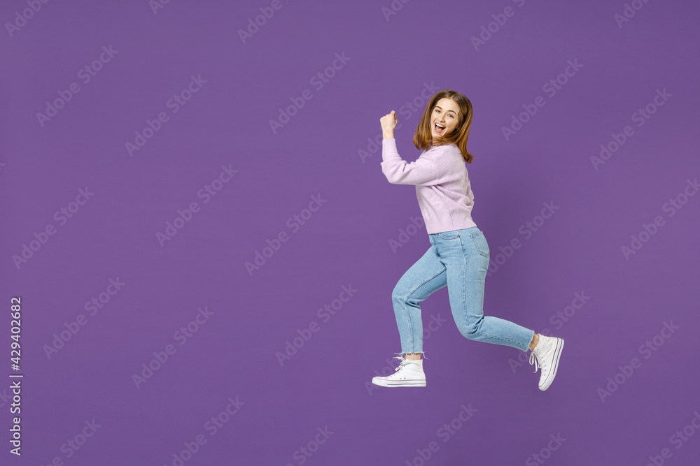 Full length side profile view of young excited smiling happy woman 20s in purple knitted sweater jump high run fast hurrying up isolated on violet background studio portrait People lifestyle concept.