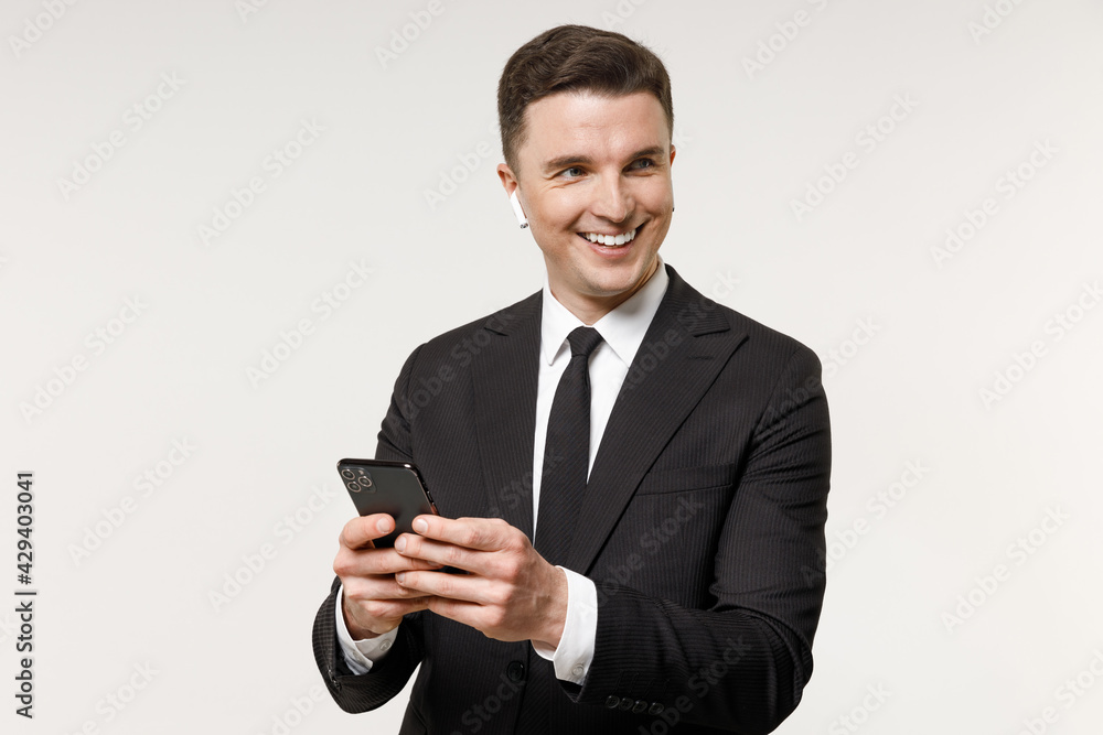 Young employee business corporate lawyer man 20s wear classic formal black grey suit shirt tie headphones air pods work in office hold mobile cell phone look aside isolated on white background studio. Young employee business corporate lawyer man 20s wear classic formal black grey suit shirt tie headphones air pods work in office hold mobile cell phone look aside isolated on white background studio.