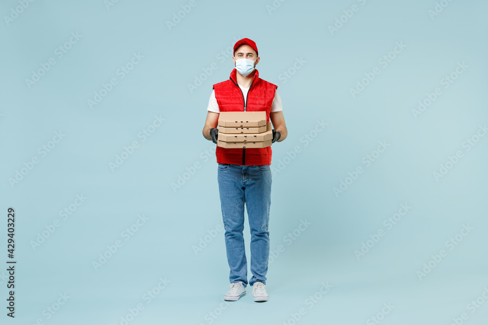 Full length delivery guy employee man in red cap t-shirt vest uniform face mask work courier service covid-19 flu virus hold pizza in cardboard flatbox run go walk isolated on pastel blue background. Full length delivery guy employee man in red cap t-shirt vest uniform face mask work courier service covid-19 flu virus hold pizza in cardboard flatbox run go walk isolated on pastel blue background.