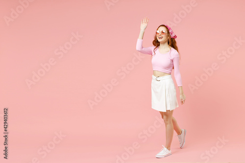 Full length side view young fun smiling happy caucasian woman in rose clothes bandana glasses walking waving hand greeeting isolated on pastel pink background studio portrait People lifestyle concept