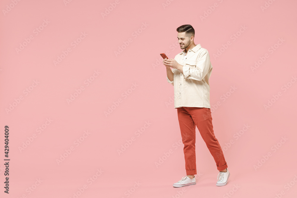 Full length young smiling happy fun fashionable friendly caucasian man in jacket white t-shirt using mobile cell phone chat online in social network walk go isolated on pastel pink background studio. Full length young smiling happy fun fashionable friendly caucasian man in jacket white t-shirt using mobile cell phone chat online in social network walk go isolated on pastel pink background studio.