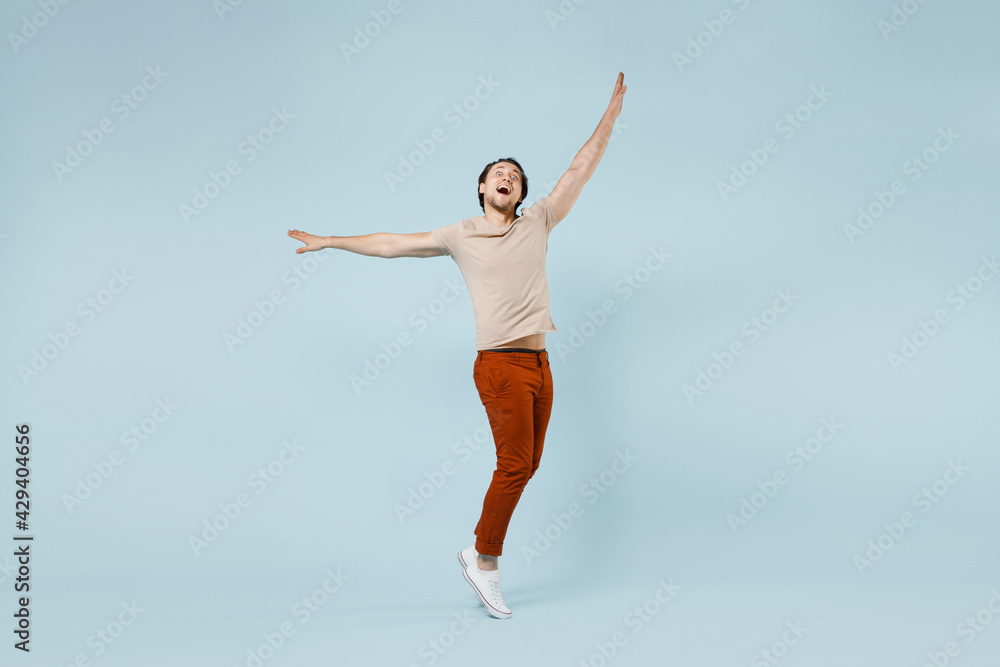 Full length young smiling happy overjoyed excited fun man 20s wear casual basic beige t-shirt standing on toes dancing with outstretched hands isolated on pastel blue color background studio portrait.