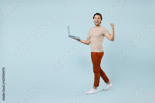 Full length side view young happy overjoyed caucasian man 20s wear casual basic beige t-shirt hold laptop pc computer do winner gesture clench fist isolated on pastel blue background studio portrait