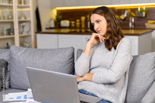 Female entrepreneur sits on the couch, works with the laptop and feels bored, 30s woman has not inspiration for freelance work, sad female feeling weary