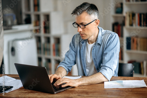 Focused smart successful gray-haired senior caucasian man wearing glasses, freelance, businessman or manager, works remotely on laptop in living room, texting with colleague or client,preparing report photo