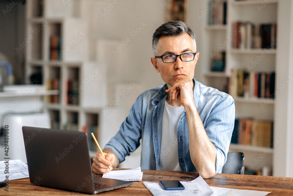 Fototapeta premium Concentrated serious senior gray-haired caucasian businessman or freelance, wearing glasses, working with laptop online, looking away, thinking over business plan, supporting his head with his hand