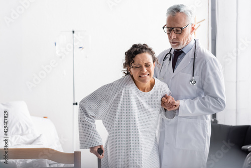 mature doctor supporting african american woman frowning from pain while walking with stick