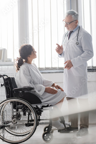 mature doctor talking to african american woman sitting in wheelchair on blurred foreground