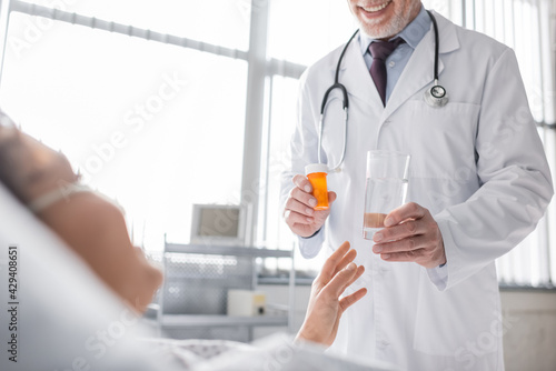 cropped view of smiling doctor holding pills and glass of water near blurred african american woman