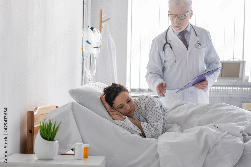 middle aged doctor with clipboard standing near upset african american woman lying in hospital bed middle aged doctor with clipboard standing near upset african american woman lying in hospital bed