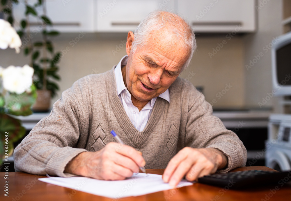 elderly pensioner writing on piece of paper at table in kitchen - obrazy, fototapety, plakaty 