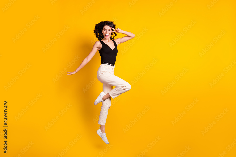 Full length body size photo of funny woman jumping up showing v-sign gesture isolated on vibrant yellow color background
