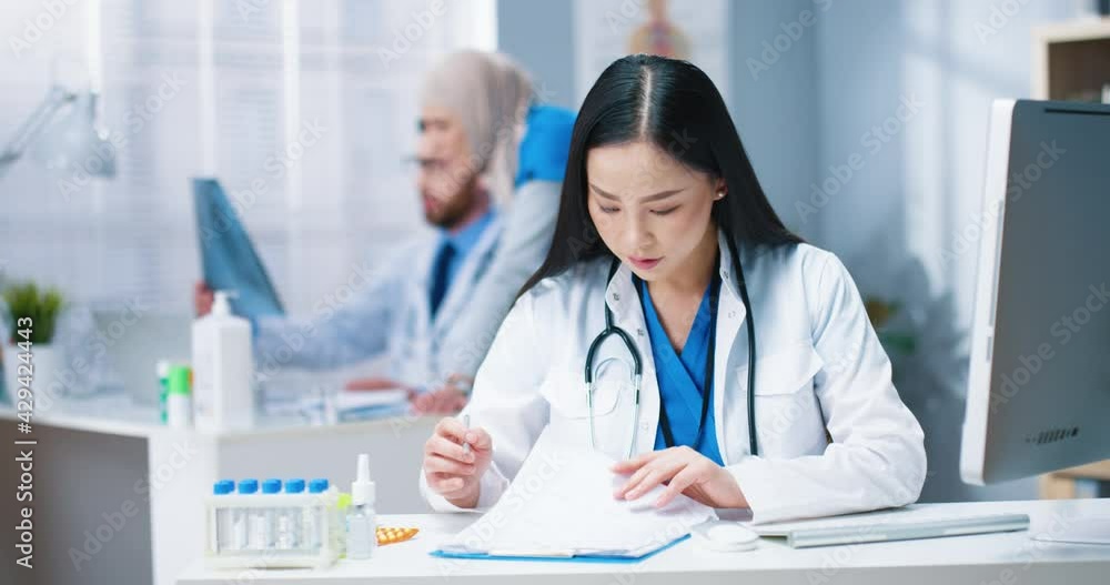 Portrait of Asian beautiful young female professional doctor in white medical coat sitting at desk at workplace in hospital cabinet and writing reading analysis results. Healthcare worker in clinic