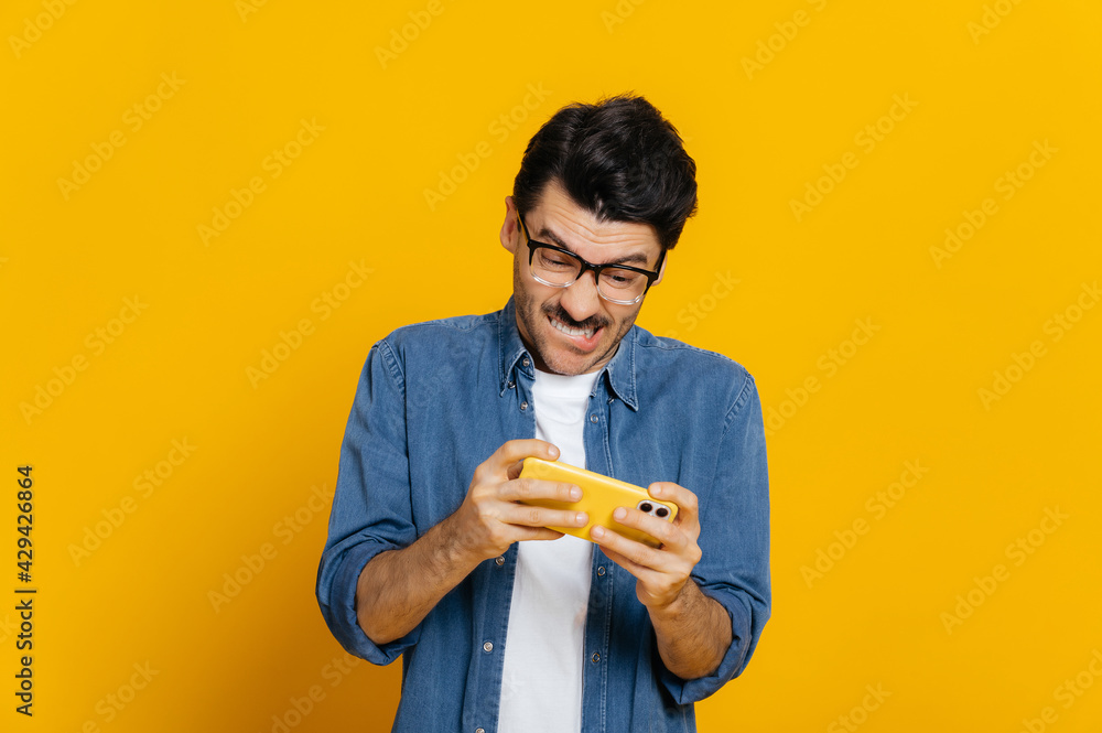 Gaming concept. Addicted caucasian male gamer with glasses, holds smartphone horizontally, plays online games, stylishly dressed, tries to win, spends free time, stands on isolated orange background Gaming concept. Addicted caucasian male gamer with glasses, holds smartphone horizontally, plays online games, stylishly dressed, tries to win, spends free time, stands on isolated orange background