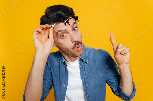 Amazed surprised caucasian guy in an orange t-shirt, shocked looking at the camera with his glasses raised, shows IDEA gesture, standing on an isolated orange background