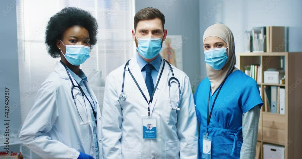 Portrait of happy young multi-ethnic male and female doctors in medical masks looking at camera in good mood at work in medical center. Team of healthcare workers in hospital. Coronavirus covid-19 Portrait of happy young multi-ethnic male and female doctors in medical masks looking at camera in good mood at work in medical center. Team of healthcare workers in hospital. Coronavirus covid-19