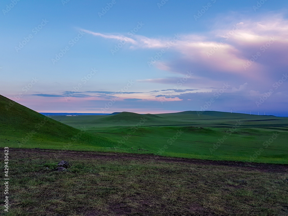 Fototapeta premium A panoramic view on a hilly landscape of Xilinhot in Inner Mongolia. Endless grassland with a few wind turbines in the back. The sun starts to set, coloring the sky pink. Thick, rainy clouds.