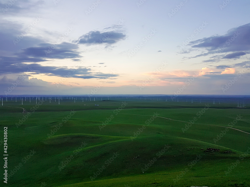 A panoramic view on a hilly landscape of Xilinhot in Inner Mongolia. Endless grassland with a few wind turbines in the back. The sun starts to set, coloring the sky orange. Thick, rainy clouds.