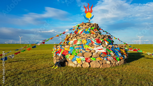 A woman in a beanie sitting on a vast pasture under a heap of stones (aobao) with colorful prayer flags attached to it. There are a lot of wind turbines in the back. Blue sky with white clouds. Spirit photo