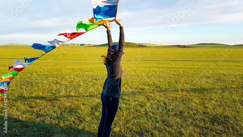 A woman holding colorful prayer flags attached to a heap of stones (aobao) above her head, while standing on a vast grassland in Xilinhot, Inner Mongolia. Endless grassland. Freedom and spirituality. photo