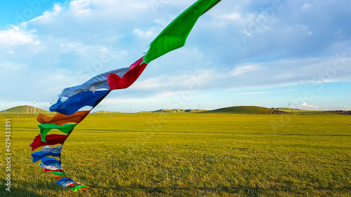 Colorful prayer flags attached to a heap of stones (aobao) on a vast grassland in Xilinhot, Inner Mongolia. A field of wind turbines in the back. Blue sky with white clouds. Spirituality