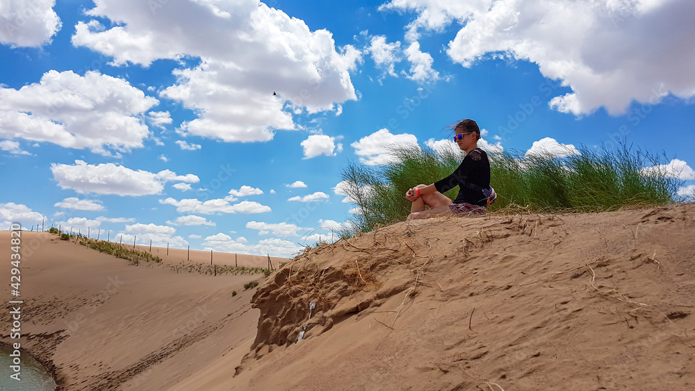 A woman in shorts sitting on a sand dune on Hunshandake Desert in the nearby of Xilinhot, Inner Mongolia. The sand dunes are overgrown with a bit of grass. Small river below the dunes. Solitude