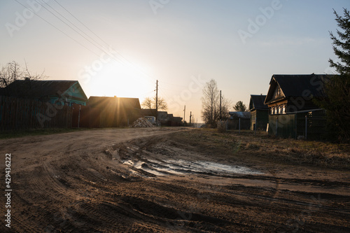 In the early spring morning in the village, the sun shines on the dirt road and wooden houses.  Muddy ground after rain
