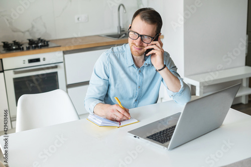 Smiling freelancer guy working from home, sits at the kitchen desk in front of the laptop, talking on the mobile phone and takes notes, multitasking man works remotely