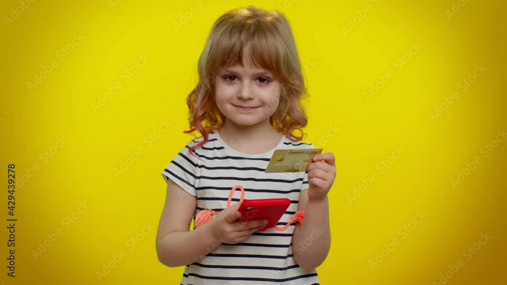 Little blonde teen kid child girl using plastic credit bank card and mobile phone while transferring money, purchases online shopping. Young children with smartphone posing on yellow studio background Little blonde teen kid child girl using plastic credit bank card and mobile phone while transferring money, purchases online shopping. Young children with smartphone posing on yellow studio background