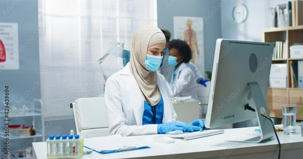 Young Arab beautiful professional female physician in medical mask working, typing and browsing online on computer sitting at workplace. Covid-19 virus, healthcare, medicine concept Young Arab beautiful professional female physician in medical mask working, typing and browsing online on computer sitting at workplace. Covid-19 virus, healthcare, medicine concept