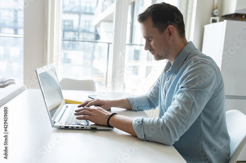 Side view on concentrated young businessman typing on the keyboard  smart man wearing smart casual shirt answering emails  messaging online on the laptop working remptely from home
