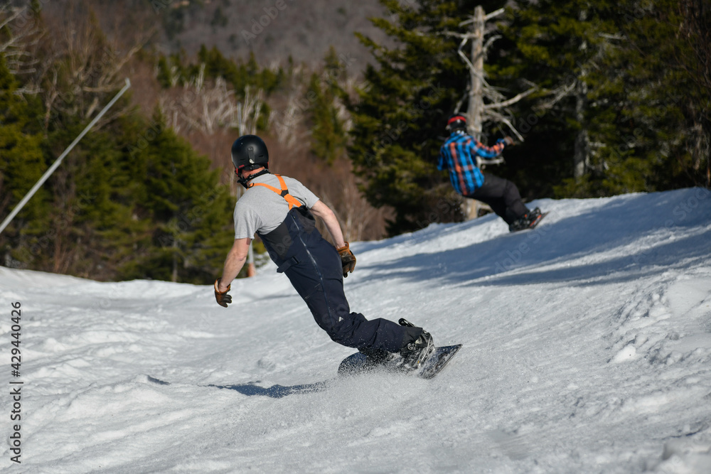 Two snowboarders seen from behind making a turn in Stowe Mountain resort in Vermont during Spring in mid-April warm sunny day.