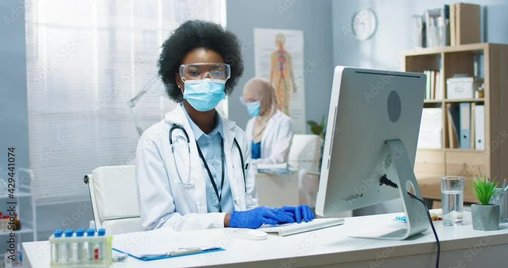 Young African American beautiful professional female physician in medical mask and goggles working, typing and browsing online on computer sitting at workplace. Covid-19 virus, medicine concept